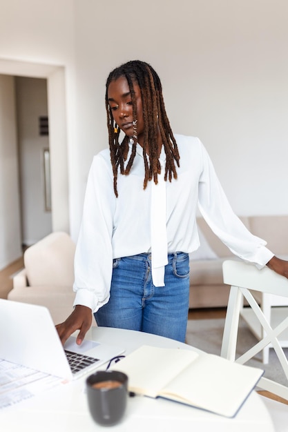 Jovem empresária afro-americana trabalhando remotamente em casa usando um laptop e desfrutando de uma xícara de café sorrindo