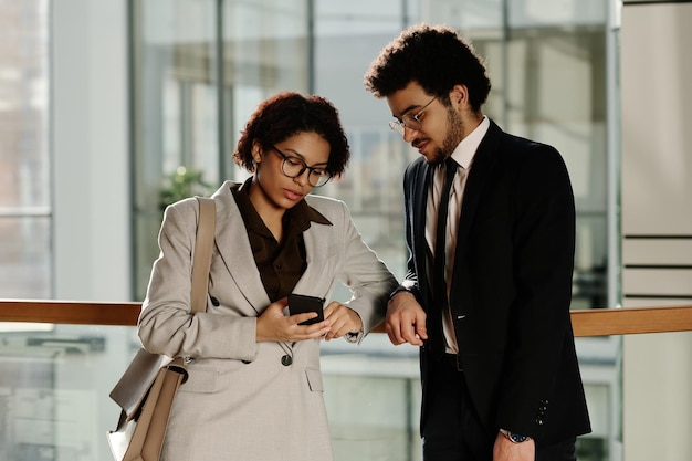 Jovem empresária apontando para smartphone e discutindo mensagem com seu colega enquanto eles stan