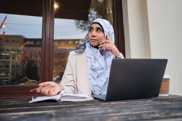 Jovem empresária árabe em hijab falando com confiança em smartphone trabalhando em laptop na rua da cidade