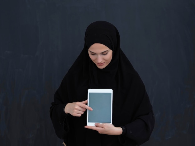 Jovem empresária árabe em roupas tradicionais ou abaya e óculos mostrando a tela do computador tablet na frente do quadro negro representando a moda e a tecnologia do Islã moderno