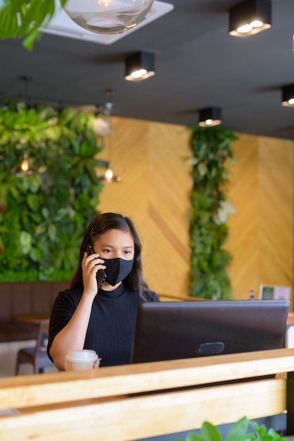 Jovem empresária asiática com máscara usando laptop e falando ao telefone dentro da cafeteria