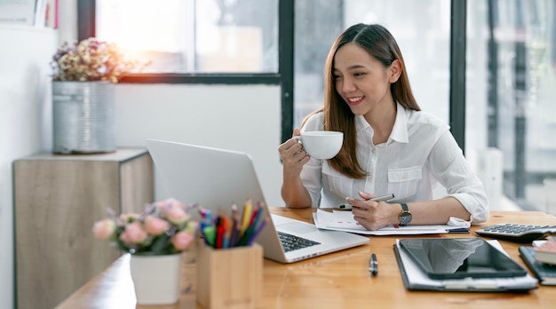 Jovem empresária asiática confiante segurando a xícara de café e trabalhando no computador portátil sentado em sua mesa de escritório