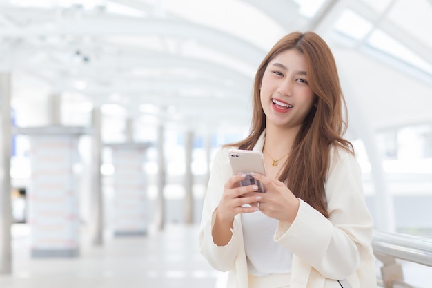 Jovem empresária asiática de terno branco sorri e segura smartphone em suas mãos em um dia de trabalho feliz