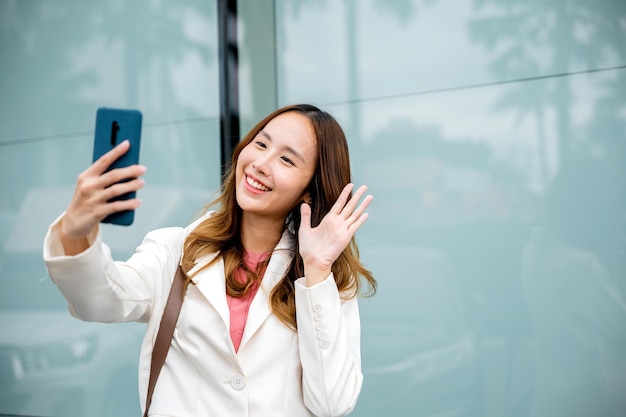 Jovem empresária asiática durante bate-papo por vídeo no telefone inteligente fora da cidade ao lado do prédio. Mulher feliz, aproveitando a luz do sol ao ar livre. Estilo de vida e tecnologia.