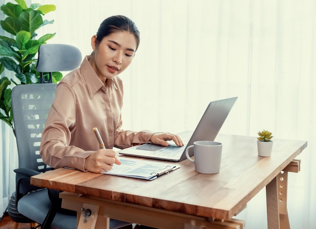 Jovem empresária asiática entusiasmada na mesa de escritório moderna usando laptop para trabalhar e escrever notas Senhora de escritório diligente e atraente trabalhando em notebook de computador em seu espaço de trabalho de escritório
