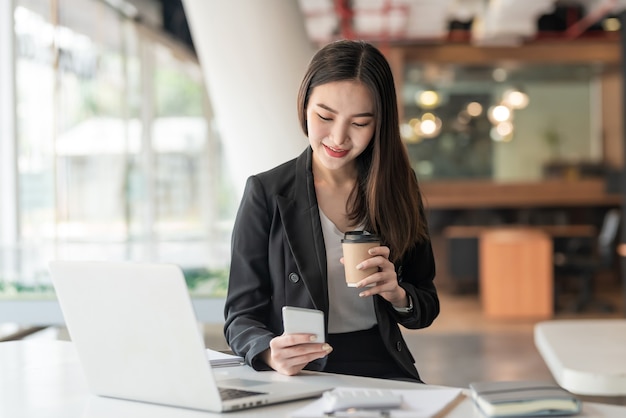 Jovem empresária asiática segurando café e usando smartphone, laptop no escritório.