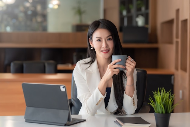 Jovem empresária asiática sentado relaxando segurando uma xícara de café e um tablet no escritório. olhando para a câmera.