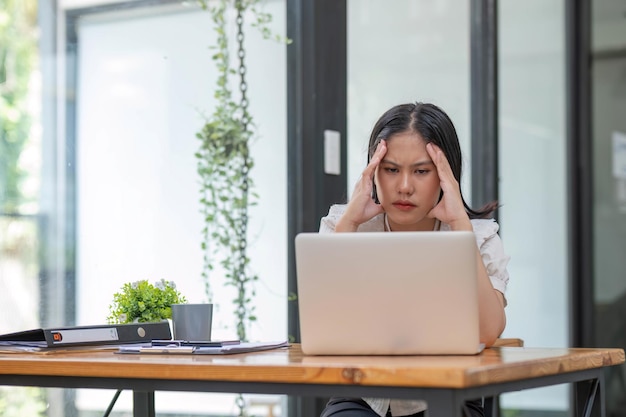 Jovem empresária asiática trabalhando em computador portátil estressada tem dor de cabeça e pensa duro do trabalho no escritório