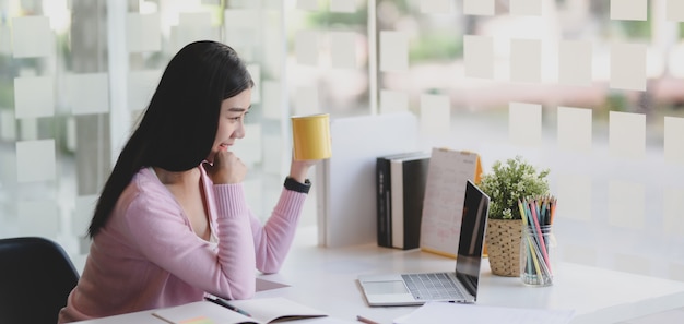 Jovem empresária asiática trabalhando em seu projeto e bebendo uma xícara de café