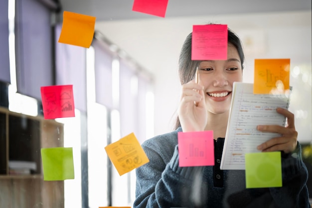 Jovem empresária atraente sorridente usando post-its na parede de vidro para escrever o plano de negócios da estratégia para o desenvolvimento crescer para o sucesso