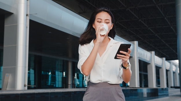 Jovem empresária bem-sucedida da ásia em roupas de escritório da moda, segurando um copo de papel descartável com uma bebida quente e usando um telefone inteligente enquanto está ao ar livre na cidade moderna urbana