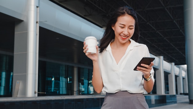 Jovem empresária bem-sucedida da Ásia em roupas de escritório da moda, segurando um copo de papel descartável com uma bebida quente e usando um telefone inteligente enquanto caminha ao ar livre na cidade moderna urbana