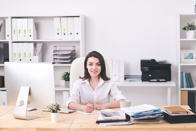 Jovem empresária bem-sucedida em trajes formais, sentada à mesa no escritório, fazendo um plano de trabalho ou anotações sobre um novo projeto de negócio