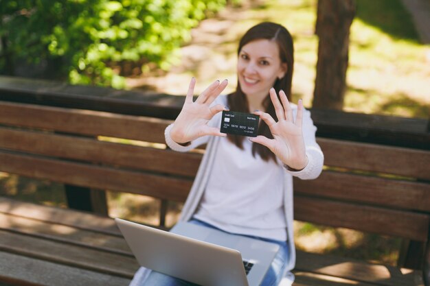 Jovem empresária bem-sucedida segurando o cartão de crédito. Mulher sentada no banco, trabalhando no computador laptop moderno no parque da cidade, na rua ao ar livre na natureza. Escritório móvel. Freelance, conceito de negócio.