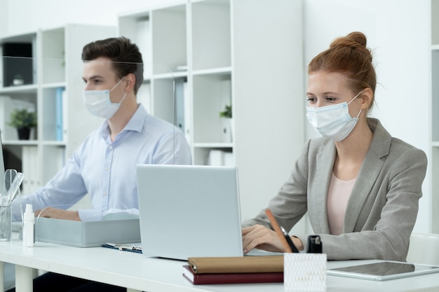 Jovem empresária bonita com máscara protetora e terno cinza olhando para a tela do laptop enquanto analisa ou insere dados contra um colega de trabalho