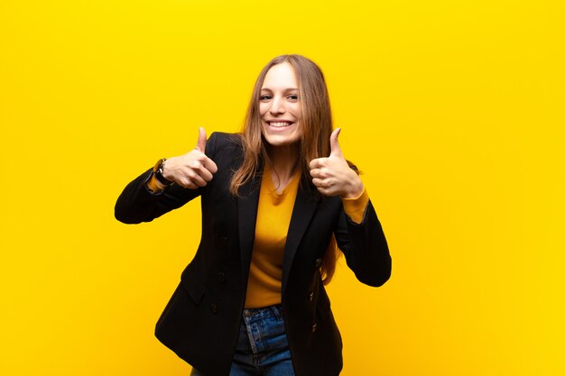 Jovem empresária bonita sorrindo amplamente olhando feliz, positivo, confiante e bem sucedido, com os dois polegares contra a parede laranja