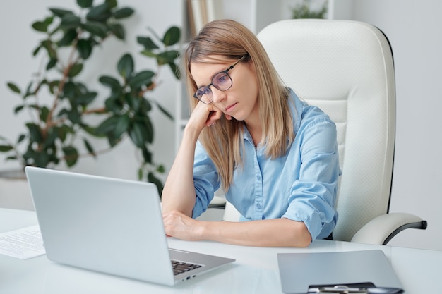 Jovem empresária cansada com longos cabelos loiros olhando tristemente para a tela do laptop enquanto analisa dados financeiros por mesa no escritório