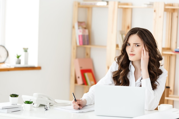 Jovem empresária cansada, sofrendo de muito tempo sentada na mesa do computador no escritório.