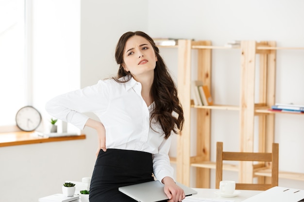 Jovem empresária cansada, sofrendo de muito tempo sentada na mesa do computador no escritório.