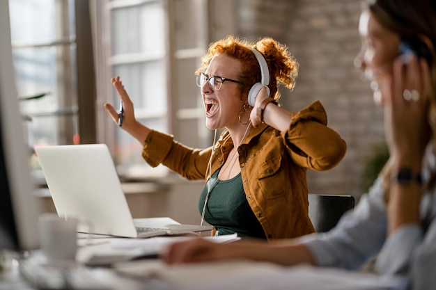 Jovem empresária casual curtindo sua música favorita em fones de ouvido e cantando enquanto trabalhava no laptop no escritório