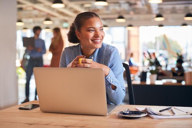 Jovem empresária com laptop sentado na mesa tomando café no escritório moderno de plano aberto