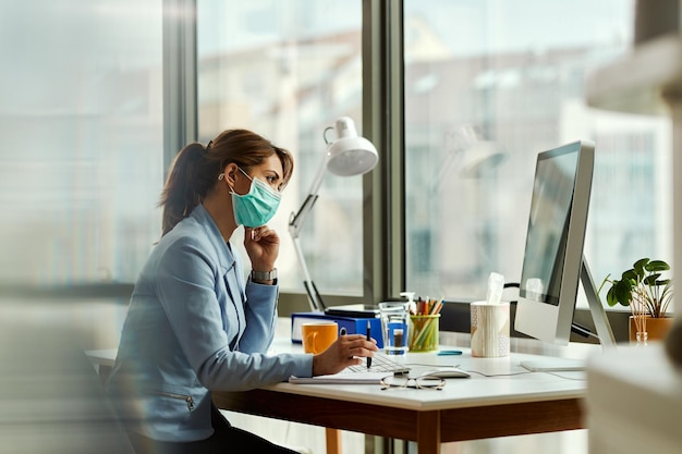 Jovem empresária com máscara facial pensando ao usar o PC desktop e trabalhando no escritório