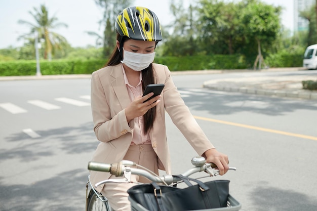 Jovem empresária com máscara protetora sentada na bicicleta e verificando as notificações no smartphone