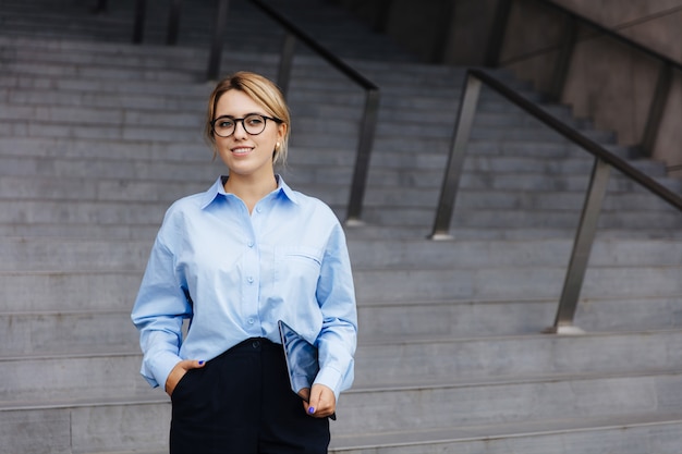 Jovem empresária com roupa formal em pé perto de prédio de escritórios com tablet digital. Conceito de pessoas, carreira de sucesso e tecnologia.