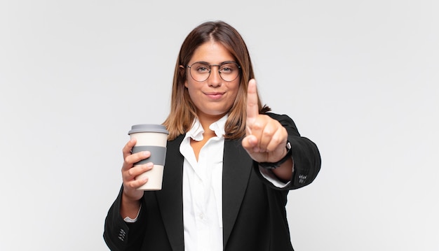 Jovem empresária com um café sorrindo com orgulho e confiança fazendo a pose número um triunfantemente, sentindo-se uma líder