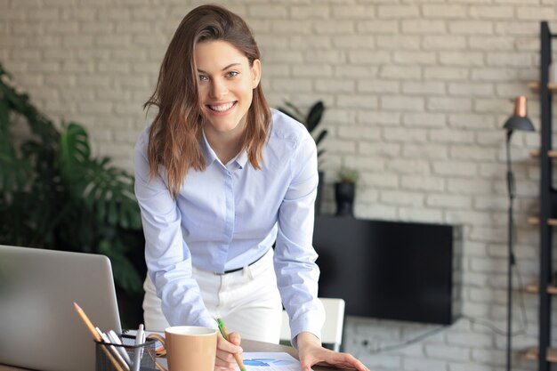 Jovem empresária confiante com um sorriso amigável atrás de sua mesa em um escritório em casa, olhando para a câmera.