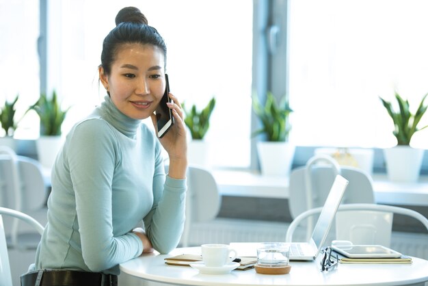 Jovem empresária confiante em roupas casuais, sentada à mesa no café, falando por smartphone e rede