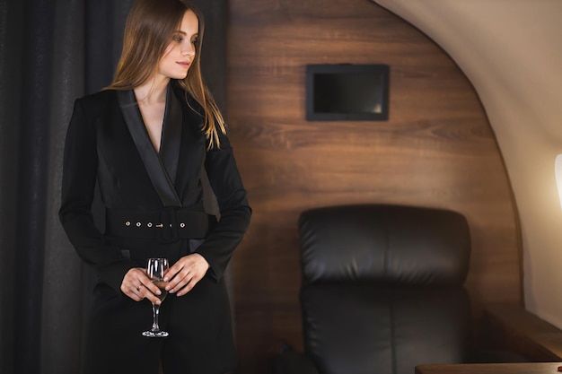 Jovem empresária confiante no jato. Uma linda garota esbelta caucasiana com cabelo comprido em um elegante vestido de jaqueta preta está de pé na cabine de um avião particular, segurando um copo de vinho na mão.