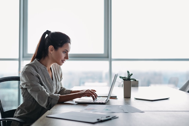 Jovem empresária de cabelos escuros vestindo camisa listrada está sentada em seu escritório e olhando para o laptop. Os documentos necessários estão sobre a mesa. Escritório moderno e luminoso.