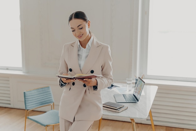 Jovem empresária de sucesso vestindo terno bege clássico, olhando no bloco de notas com lista de tarefas ou plano de negócios em pé no armário de escritório perto do local de trabalho. Feliz dia de trabalho de planejamento feminino