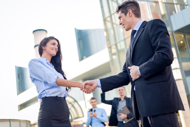 Jovem empresária e empresário apertando as mãos na frente da empresa enquanto no fundo em pé deles da equipe de negócios.