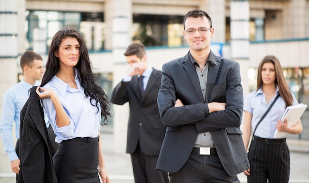 Jovem empresária e empresário, com sua equipe em segundo plano, em frente ao prédio de escritórios, com um sorriso olhando para a câmera.