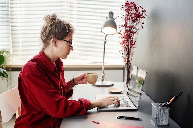 Jovem empresária em óculos, sentada à mesa, bebendo café e trabalhando online no laptop em casa