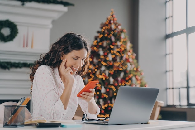 Jovem empresária espanhola feliz usando smartphone enquanto trabalhava laptop em casa no Natal