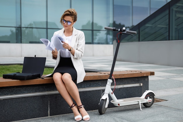 Jovem empresária está sentada no banco com vários documentos em papel e uma scooter elétrica ao lado dela