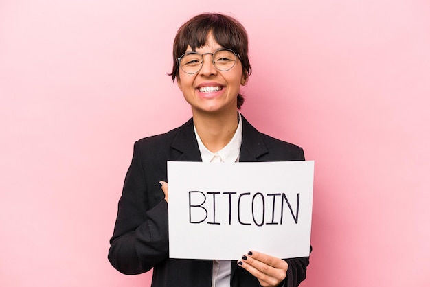 Jovem empresária hispânica segurando um cartaz de bitcoin isolado em fundo rosa rindo e se divertindo