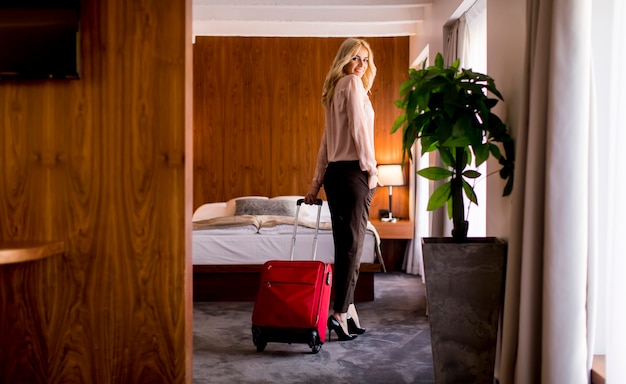 Foto jovem empresária loira chega em um quarto de hotel com mala vermelha