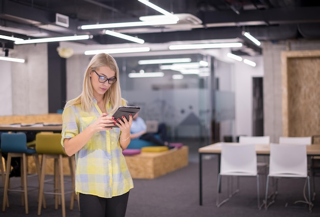 jovem empresária loira trabalhando online usando tablet digital em pé no escritório de inicialização moderno com equipe de negócios multiétnica em segundo plano