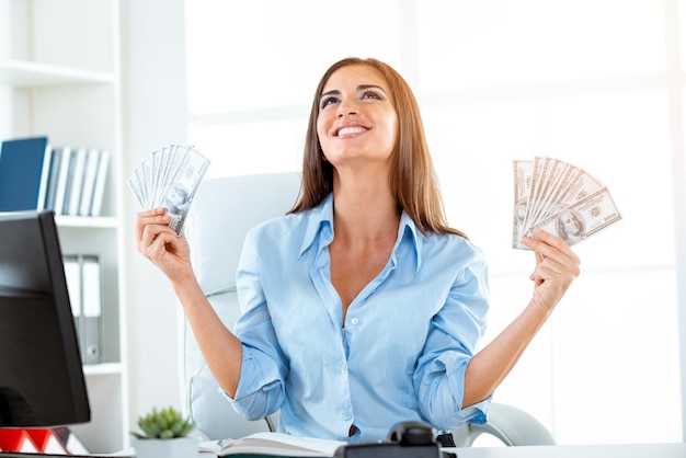 Foto jovem empresária no escritório, sentado na mesa do escritório, segurando dinheiro e com uma expressão de felicidade no rosto olhando para cima.