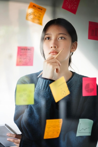 Jovem empresária pensando e planejando estratégia de negócios usando notas post-it na parede de vidro para o desenvolvimento crescer para o sucesso