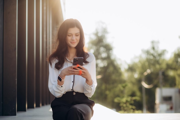 Jovem empresária positiva em óculos com xícara de café na mão usando telefone celular enquanto está de pé na rua ensolarada perto do prédio de escritórios conversando on-line com amigos durante o intervalo no trabalho