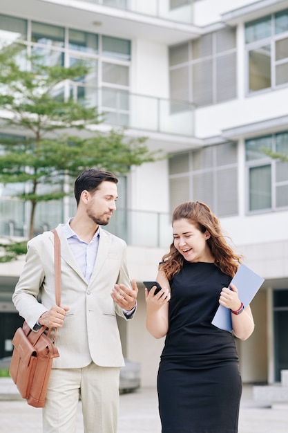 Jovem empresária positiva lendo mensagem de texto no smartphone ao caminhar ao ar livre e discutir um novo projeto criativo com um colega