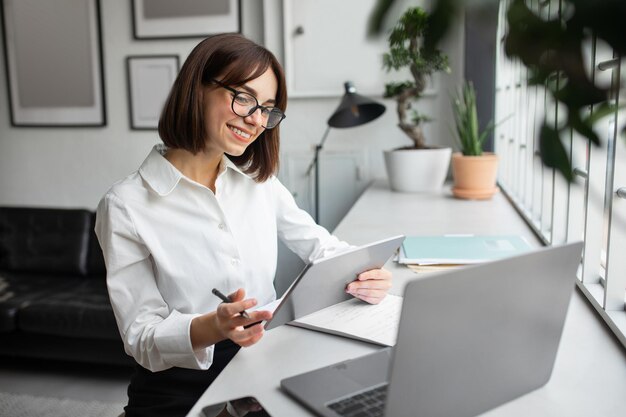 Jovem empresária positiva trabalhando com tablet digital e laptop sentada em uma mesa em um escritório aconchegante