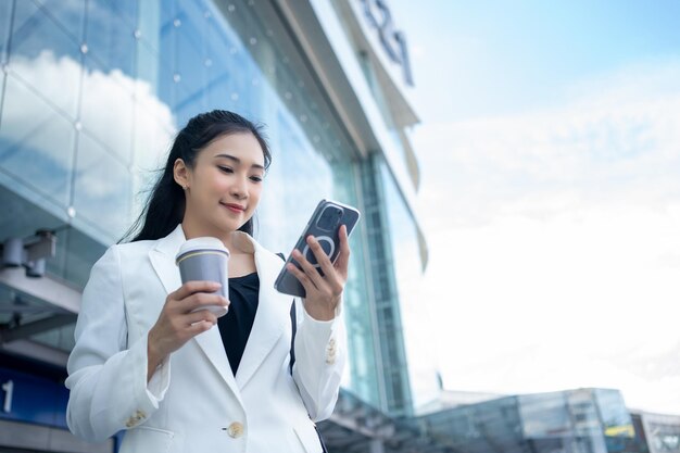 Foto jovem empresária segurando uma xícara de café quente e usando um smartphone enquanto caminha pela cidade