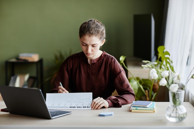 Jovem empresária sentada à mesa com o laptop e usando o calendário para planejar seu trabalho em casa