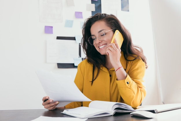 Jovem empresária sorridente de óculos, sentada no local de trabalho Garota vestindo camisa amarela lendo papel e discutindo com o cliente no telefone